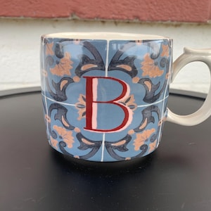 May include: Ceramic mug with a blue tile pattern and the letter "B" in red. The mug has a white handle and a light blue background. The design includes orange and dark blue floral elements.