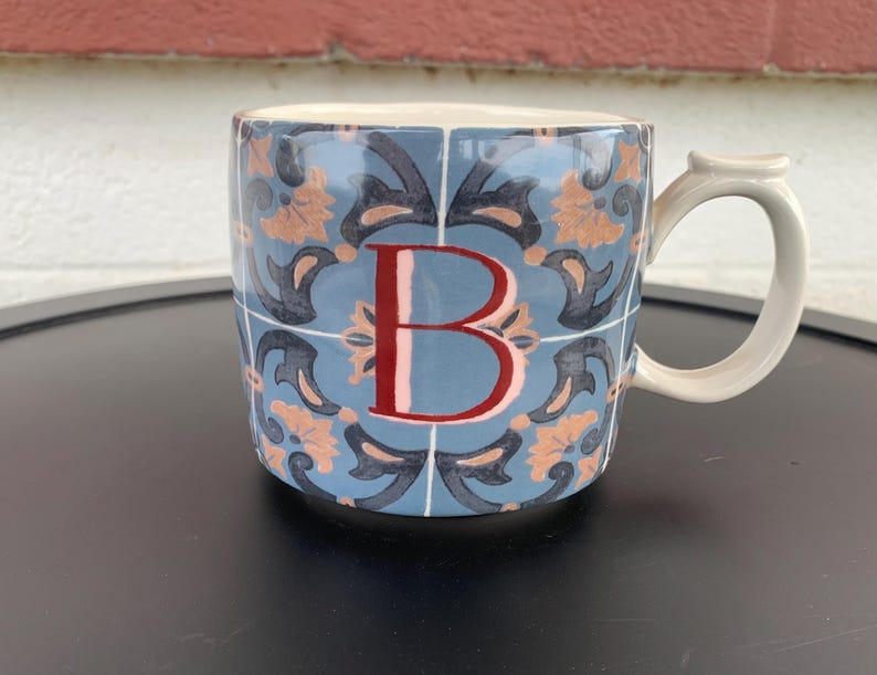 May include: Ceramic mug with a blue tile pattern and the letter "B" in red. The mug has a white handle and a light blue background. The design includes orange and dark blue floral elements.