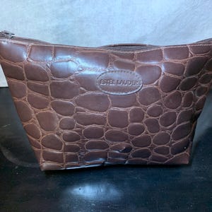 May include: Brown cosmetic bag with a crocodile skin texture. The bag has a zipper closure and a small oval patch with the brand name "Estee Lauder" embossed on it. The bag is made of a dark brown material.