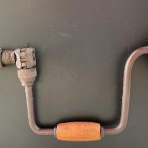 May include: A vintage hand drill with a wooden handle and a chuck for holding drill bits. The drill has a curved metal frame and a wooden knob on the opposite end of the chuck. The drill is made of metal and wood.