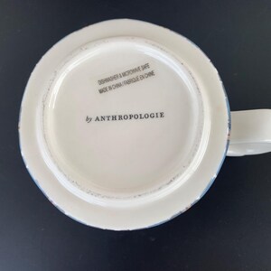 May include: White ceramic mug with a handle, featuring a distressed blue and brown rim. The bottom of the mug displays the text "DISHWASHER & MICROWAVE SAFE MADE IN CHINA / FABRIQUE EN CHINE" and "by ANTHROPOLOGIE".