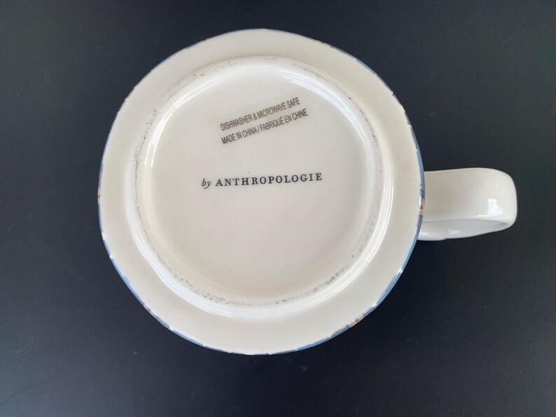 May include: White ceramic mug with a handle, featuring a distressed blue and brown rim. The bottom of the mug displays the text "DISHWASHER & MICROWAVE SAFE MADE IN CHINA / FABRIQUE EN CHINE" and "by ANTHROPOLOGIE".