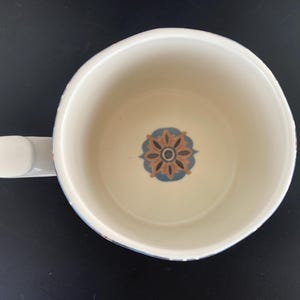 May include: Overhead view of a white ceramic mug with a handle. The interior features a floral design in brown, blue, and cream. The mug sits on a black surface.