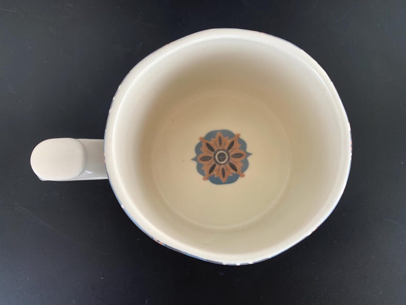 May include: Overhead view of a white ceramic mug with a handle. The interior features a floral design in brown, blue, and cream. The mug sits on a black surface.
