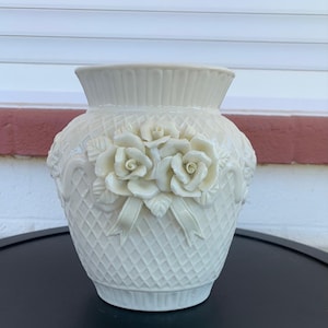 May include: Cream-colored ceramic vase with a textured diamond pattern and three-dimensional rose embellishments. The vase has a wide opening and a decorative ribbon design. A classic piece for floral arrangements.