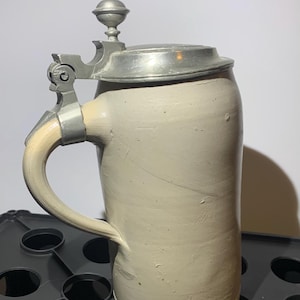 May include: A tall, cylindrical stoneware beer stein with a light beige, matte finish. The vessel features a sturdy, curved handle and a hinged, metallic pewter-colored lid topped with a small, decorative finial knob.