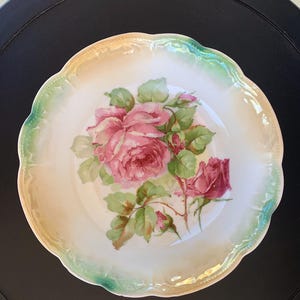 May include: A round porcelain plate featuring a central illustration of pink and deep red roses with green leaves. The plate has a scalloped edge with a soft, iridescent green and cream-colored border, creating a delicate and decorative appearance.