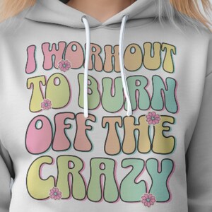 Op de afbeelding: Een lichtgrijze hoodie met een retro-grafische afbeelding met de tekst "I workout to burn off the crazy". De afbeelding is in tinten roze, geel, groen en blauw met kleine bloem accenten.