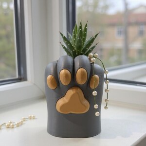 May include: A grey cat paw-shaped planter with tan paw pads, holding a succulent plant. A string of pearls dangles from the planter. The planter is on a white windowsill.