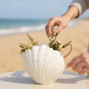 May include: A white ceramic seashell-shaped vase filled with greenery and small white flowers. The vase is on a white table, with a beach and ocean in the background. A person's hands are near the vase.