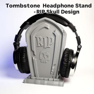 May include: A grey tombstone-shaped headphone stand with a skull design and the letters "RIP". Black headphones rest on the stand. The text "Tombstone Headphone Stand - RIP Skull Design" is at the top.