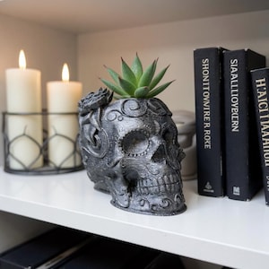 May include: A dark grey skull-shaped planter with a succulent plant, decorated with floral patterns. It sits on a white shelf with lit candles and black books. The planter is a unique home decor item.