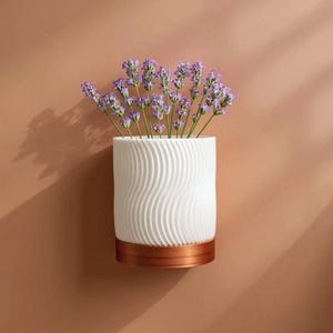 May include: A white ceramic vase with a wavy pattern, containing purple lavender flowers. The vase features a copper-coloured base and is set against a warm brown backdrop. A decorative floral arrangement.