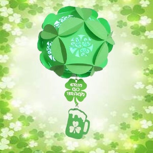 Puede incluir: Una linterna de papel verde con diseño de trébol, perfecta para el Día de San Patricio. La linterna está adornada con un dije en forma de trébol que dice "Erin Go Bragh" y un dije de jarra de cerveza con un trébol. El fondo es verde suave con acentos de trébol.
