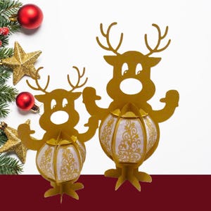 May include: Two gold-colored reindeer-shaped lanterns with white and gold patterned globes. The lanterns are on a white background with Christmas decorations, including a fir branch, red berries, gold stars, and red ornaments.