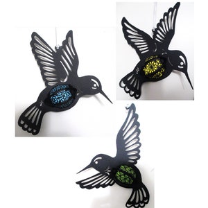 Könnte beinhalten: Schwarzes Kolibri-Ornament mit aufwendig ausgeschnittenen Flügeln und Körper. Der Körper des Kolibris zeigt ein dekoratives Ei mit blauem, gelbem oder grünem Muster. Das Ornament hängt an einer weißen Schnur, ideal für die Inneneinrichtung.