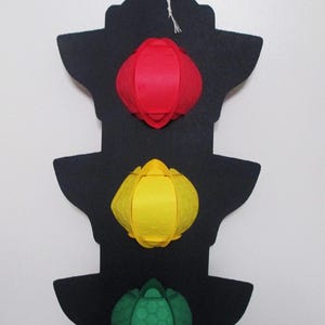 May include: A black felt traffic light decoration with three colourful paper lanterns. The top lantern is red, the middle is yellow, and the bottom is green. The decoration hangs from a white string.