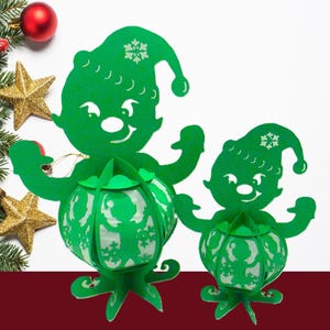 May include: Two green paper elf lanterns with snowflake details, each holding a round lantern. The lanterns are on a white background with Christmas decorations, including a red ornament, gold stars, and evergreen branches.