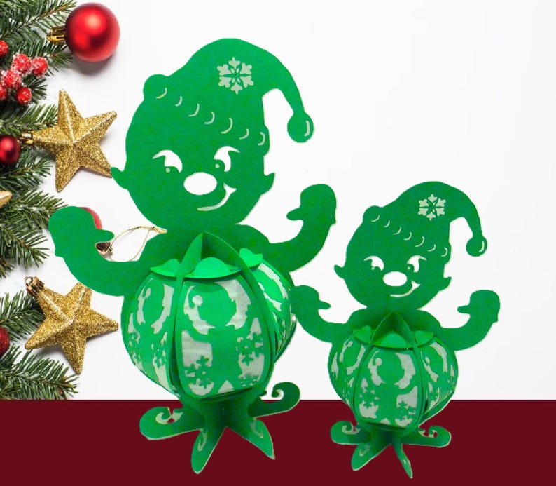 May include: Two green paper elf lanterns with snowflake details, each holding a round lantern. The lanterns are on a white background with Christmas decorations, including a red ornament, gold stars, and evergreen branches.
