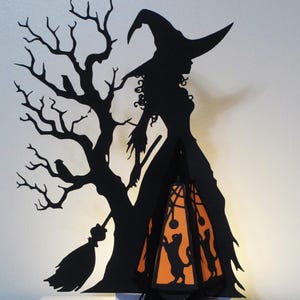 May include: A black silhouette of a witch with a pointed hat, standing next to a bare tree with a broom. An orange lantern with black cat cutouts hangs from the witch's hand. The Halloween-themed decoration is set against a white background.