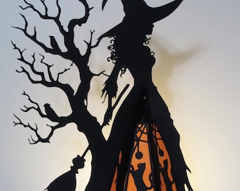 Puede incluir: Silueta negra de una bruja con sombrero puntiagudo, junto a un árbol desnudo con una escoba. Una linterna naranja con recortes de gatos negros cuelga de la mano de la bruja. Decoración de Halloween sobre fondo blanco.