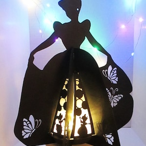 May include: A black silhouette of a woman in a ball gown, with butterfly cutouts on the skirt. The interior has a warm light, and the design is reminiscent of a fairy tale. The base is a light colour.