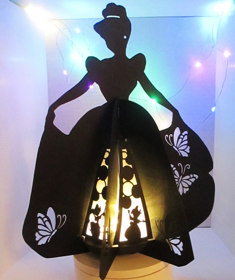 May include: A black silhouette of a woman in a ball gown, with butterfly cutouts on the skirt. The interior has a warm light, and the design is reminiscent of a fairy tale. The base is a light colour.