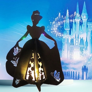 May include: A black silhouette of a woman in a ball gown, with butterfly cutouts and a glowing interior. The background features a castle and a starry sky. The lantern is a decorative item.