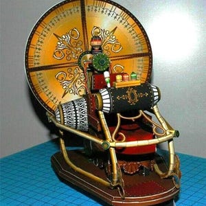 May include: A detailed paper model of a time machine featuring a large circular golden clock face with ornate patterns, a central seat, and a black cylindrical console with three small colored lights. The structure has gold-toned railings.