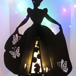 May include: A black silhouette of a woman in a ball gown, with butterfly cutouts on the skirt. The interior has a warm glow, with a silhouette of a mouse. Decorative fairy lights are in the background.
