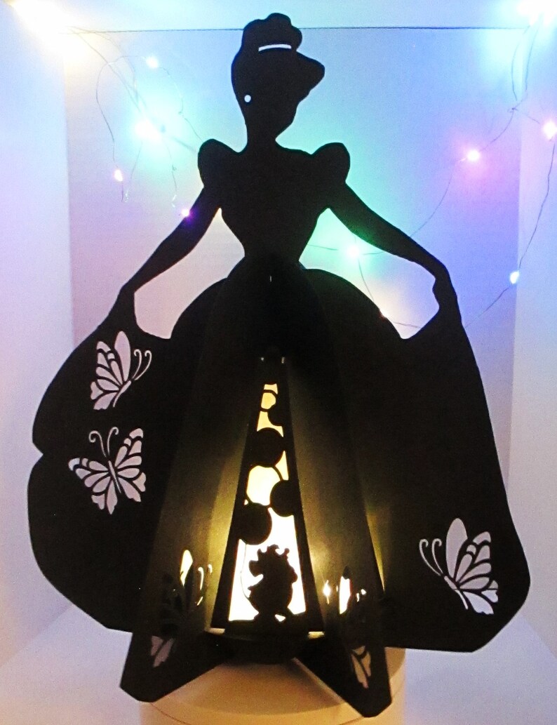 May include: A black silhouette of a woman in a ball gown, with butterfly cutouts on the skirt. The interior has a warm glow, with a silhouette of a mouse. Decorative fairy lights are in the background.