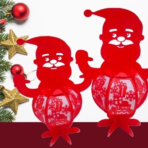 May include: Two red Santa Claus lanterns with a festive design. The lanterns have a round, patterned body and a Santa Claus head. Christmas decorations, including ornaments and stars, are in the background.