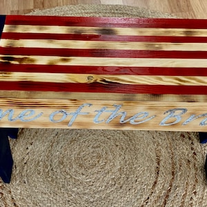 May include: A wooden bench with a distressed American flag design. The bench is painted blue and has the words "Home of the Brave" inscribed on the front.