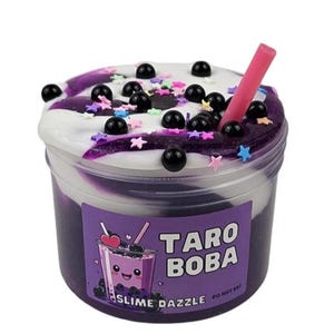 Peut inclure: Slime violet et blanc dans un récipient transparent avec une paille rose. Le slime est surmonté de billes de boba noires et de vermicelles colorés. Le récipient porte une étiquette indiquant "Taro Boba Slime Dazzle" et "Ne pas manger".