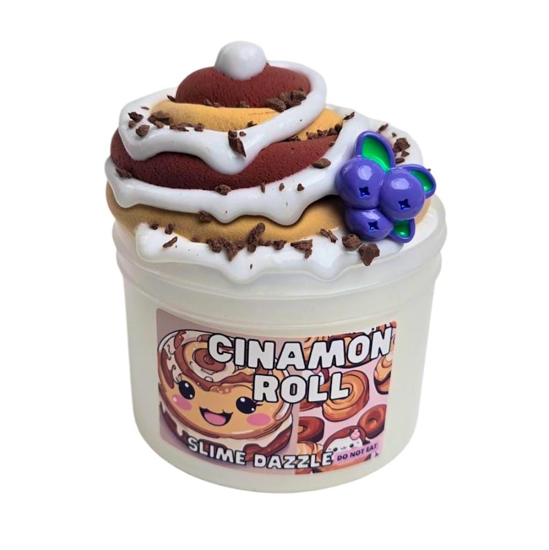May include: A clear plastic container filled with a brown, white, and tan slime that resembles a cinnamon roll. The slime is topped with a white frosting and a purple blueberry. The container has a label that reads "Cinnamon Roll Slime Dazzle" with a cartoon cinnamon roll illustration.
