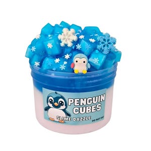 May include: A clear plastic container filled with blue slime, decorated with snowflake accents and a small penguin figurine. The container has a blue and white color scheme, with the words "Penguin Cubes Slime Dazzle" printed on the front.