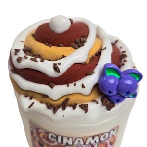 May include: A white container of slime with a cinnamon roll design. The slime is topped with white frosting and chocolate sprinkles. The container has a purple and green charm attached to it.