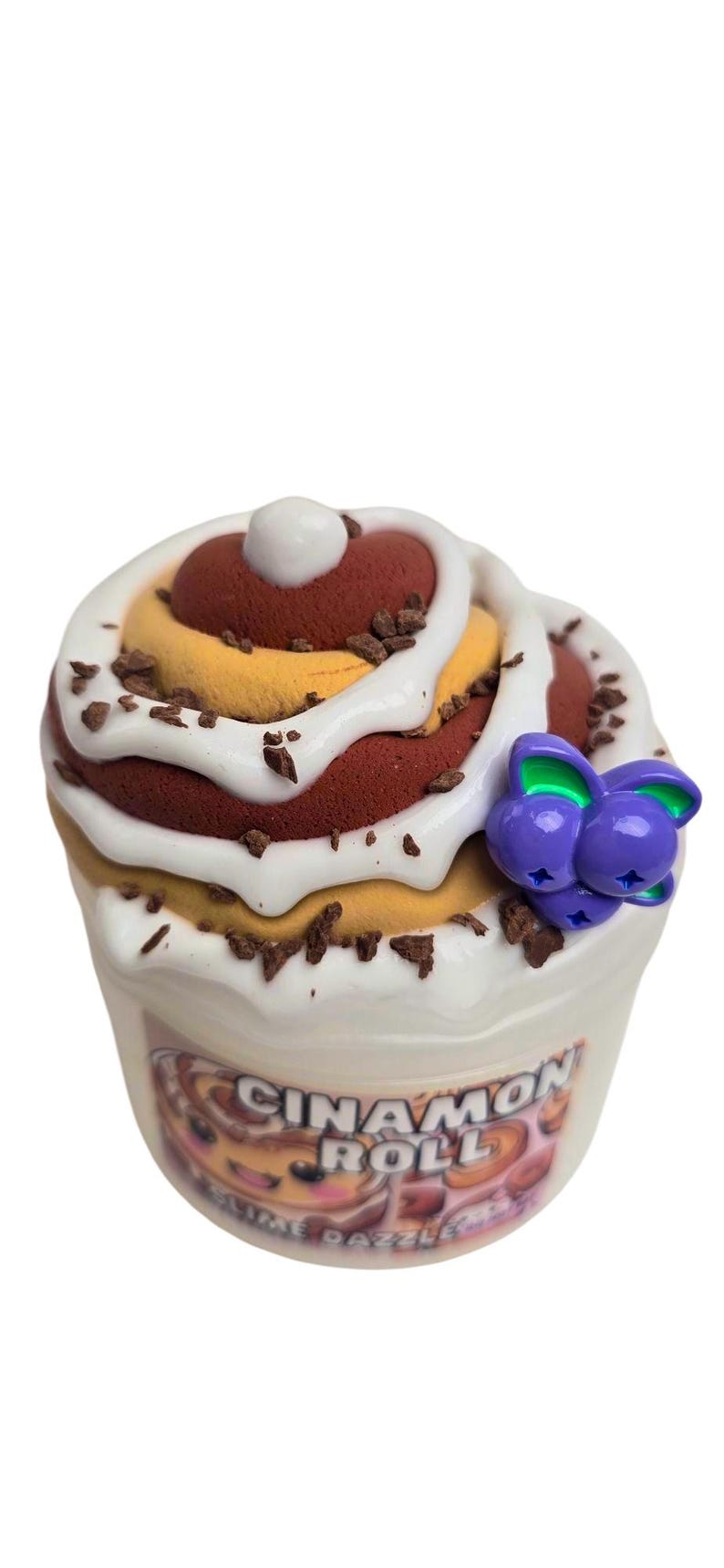 May include: A white container of slime with a cinnamon roll design. The slime is topped with white frosting and chocolate sprinkles. The container has a purple and green charm attached to it.
