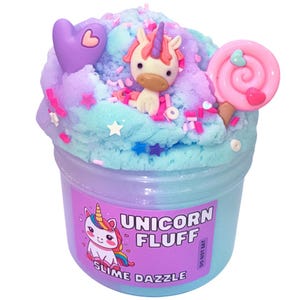 Puede incluir: Un recipiente de plástico transparente lleno de slime azul, morado y rosa, coronado con una figura de unicornio, una piruleta rosa y un corazón morado. El recipiente tiene una etiqueta que dice "Unicorn Fluff Slime Dazzle."