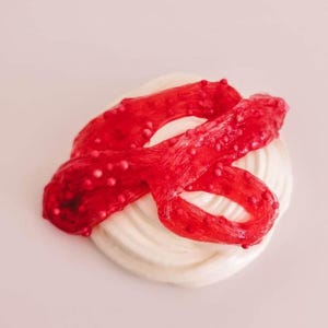 May include: A swirl of white, creamy substance is topped with a vibrant red, ribbon-like material dotted with small, pinkish-red beads. The background is a soft, pale pink.