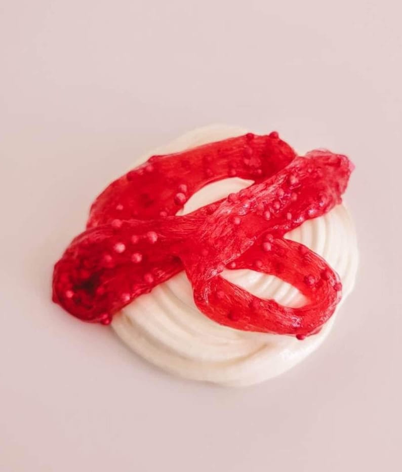 May include: A swirl of white, creamy substance is topped with a vibrant red, ribbon-like material dotted with small, pinkish-red beads. The background is a soft, pale pink.