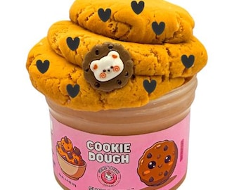 May include: A clear plastic jar filled with tan slime topped with two golden-brown cookie-shaped pieces. The jar features a pink label with the text COOKIE DOUGH, SLIME DAZZLE, and illustrations of a cookie bowl and a smiling chocolate chip cookie.