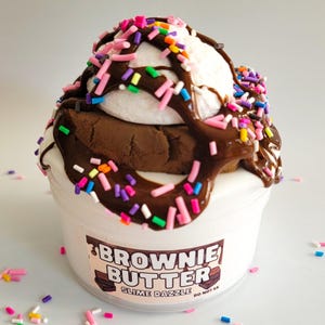 Könnte beinhalten: Ein weißes Glas Slime mit einem braunen Brownie und einer weißen Marshmallow-Füllung. Der Slime ist mit Schokoladensauce und bunten Streuseln bedeckt. Das Glas hat ein Etikett mit der Aufschrift "Brownie Butter Slime Dazzle" und "Nicht essen".