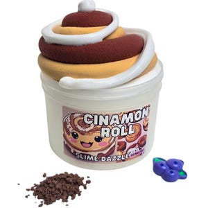 May include: A white plastic container with a clear lid filled with brown and white slime. The container has a label that says "Cinnamon Roll" and "Slime Dazzle". There is a small pile of brown sprinkles and a purple blueberry charm next to the container.