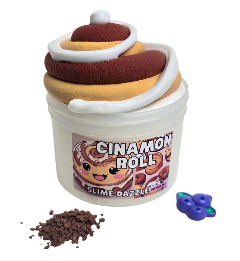 May include: A white plastic container with a clear lid filled with brown and white slime. The container has a label that says "Cinnamon Roll" and "Slime Dazzle". There is a small pile of brown sprinkles and a purple blueberry charm next to the container.