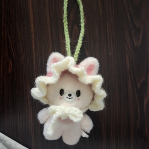 May include: A plush, light pink bear-shaped keychain with a white and pink floral hat and bow. The bear has a small black nose and eyes. A green crocheted loop is attached for hanging. The background is a dark brown wooden surface.