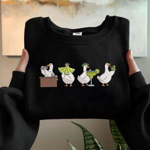 Puede incluir: Sudadera negra con una ilustraci&oacute;n caprichosa de cuatro gansos blancos. Cada ganso se representa en una situaci&oacute;n diferente, incluso detr&aacute;s de una barra, con una capa, con una margarita y sosteniendo una bebida. Los gansos llevan sombreros verdes.