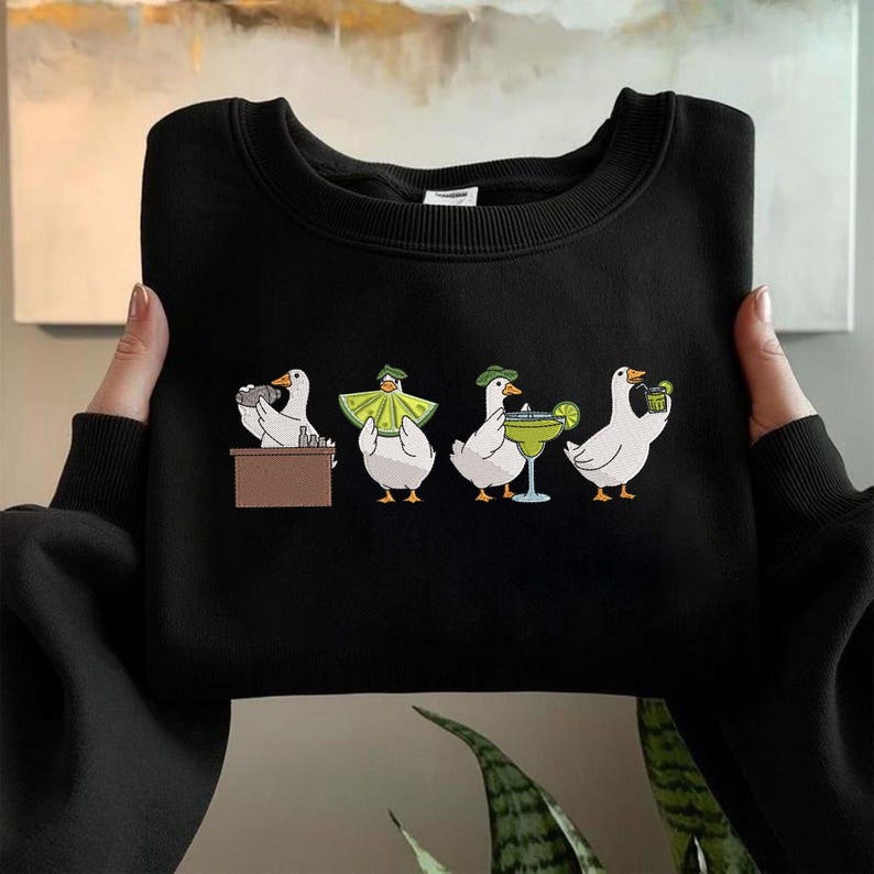 Puede incluir: Sudadera negra con una ilustraci&oacute;n caprichosa de cuatro gansos blancos. Cada ganso se representa en una situaci&oacute;n diferente, incluso detr&aacute;s de una barra, con una capa, con una margarita y sosteniendo una bebida. Los gansos llevan sombreros verdes.