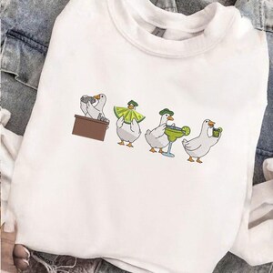 Puede incluir: Sudadera blanca con un dise&ntilde;o bordado caprichoso de gansos. Los gansos se representan en varias poses, incluyendo uno detr&aacute;s de una barra, uno con un sombrero de lima y otros con c&oacute;cteles. El dise&ntilde;o es en tonos blancos, verdes y marrones.