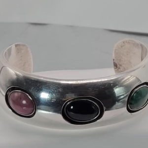 May include: Silver cuff bracelet with four oval-shaped gemstones. The stones are black, pink, green, and a darker green.